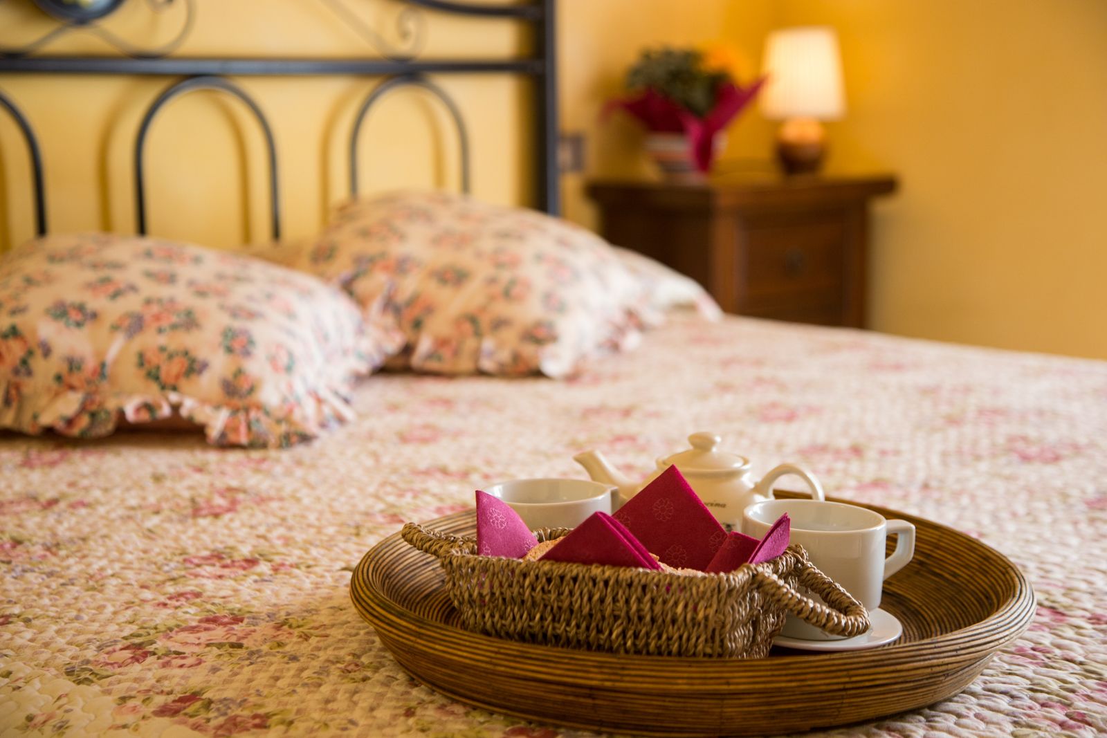 Een bed met een bloemenprint, een rieten dienblad met theekoppen en een theepot, en een nachtkastje met een lamp en een plant.