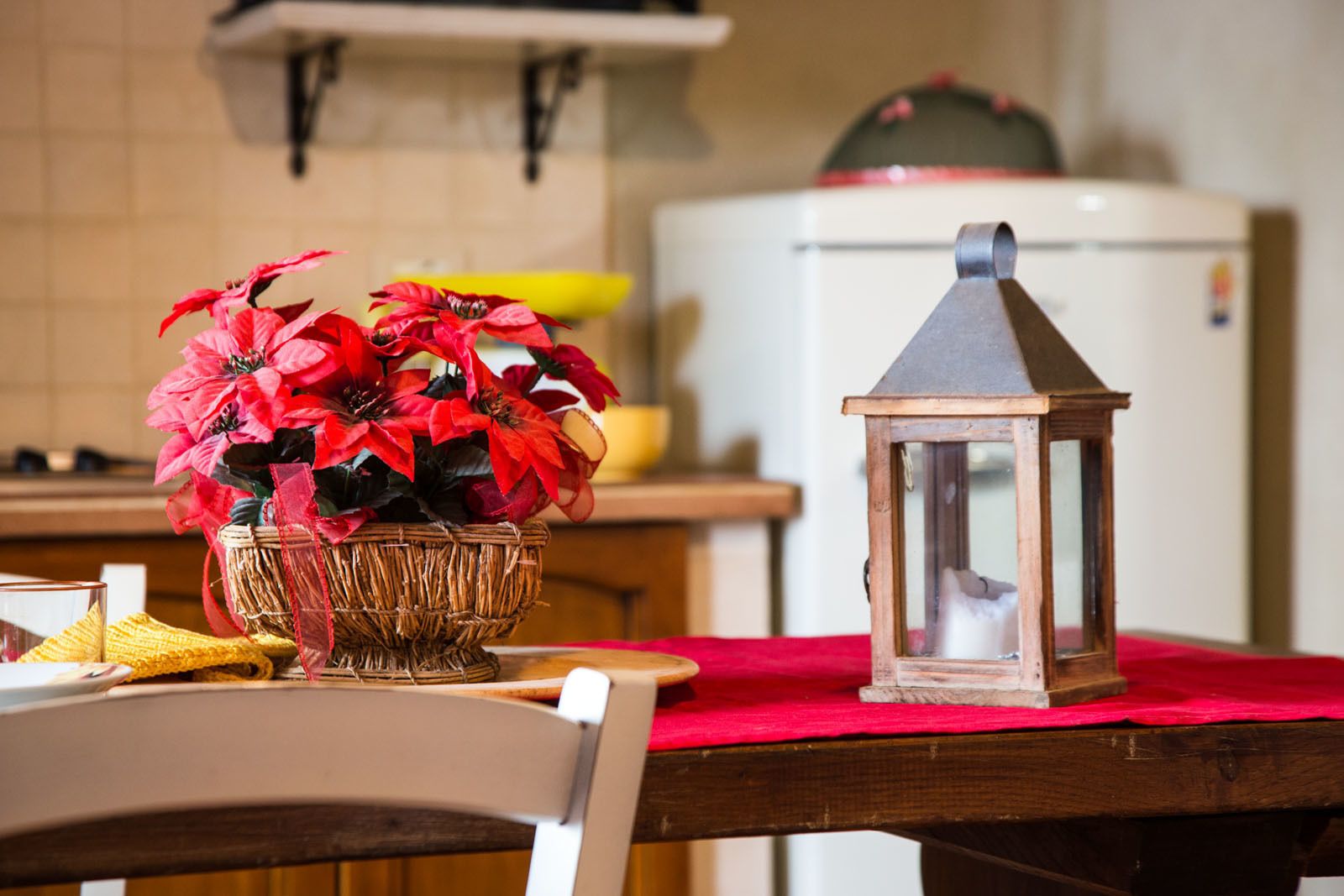A kitchen with a wicker basket containing red flowers, a wooden table with a red cloth, and a lantern.