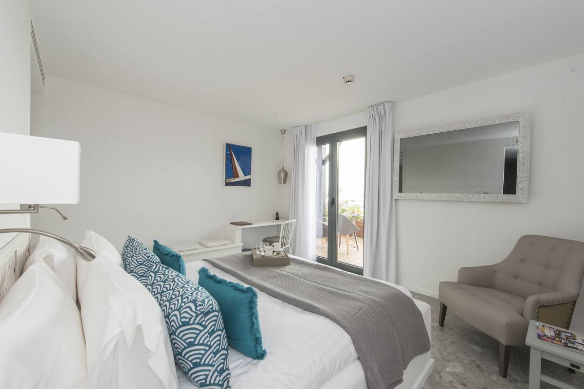 Quarto de hotel com cama, cadeira, escrivaninha e uma janela grande.