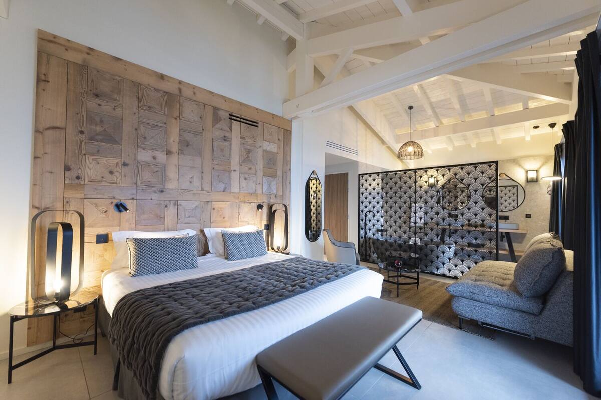 Quarto moderno com uma cama grande, mesas de cabeceira, um banquinho e um espaço de estar com parede estampada e espelho.