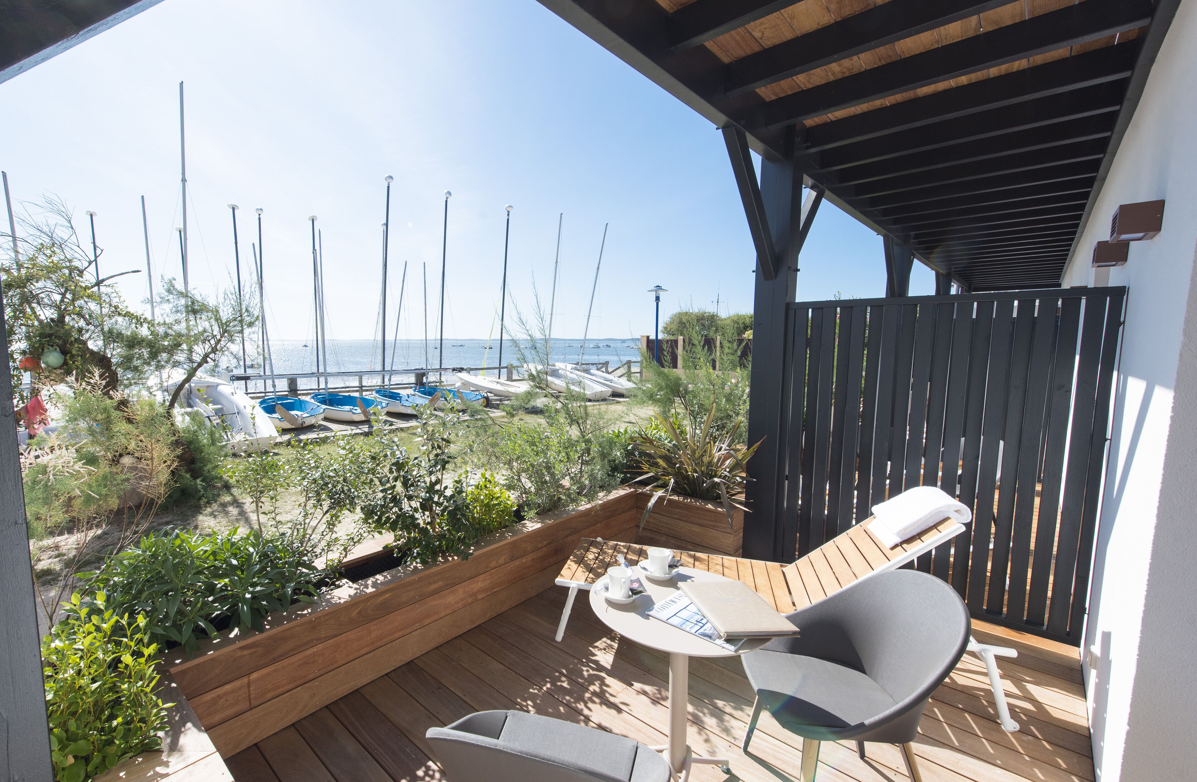 Terraza de madera con una mesa y dos sillas, con vistas a un puerto deportivo y barcos.