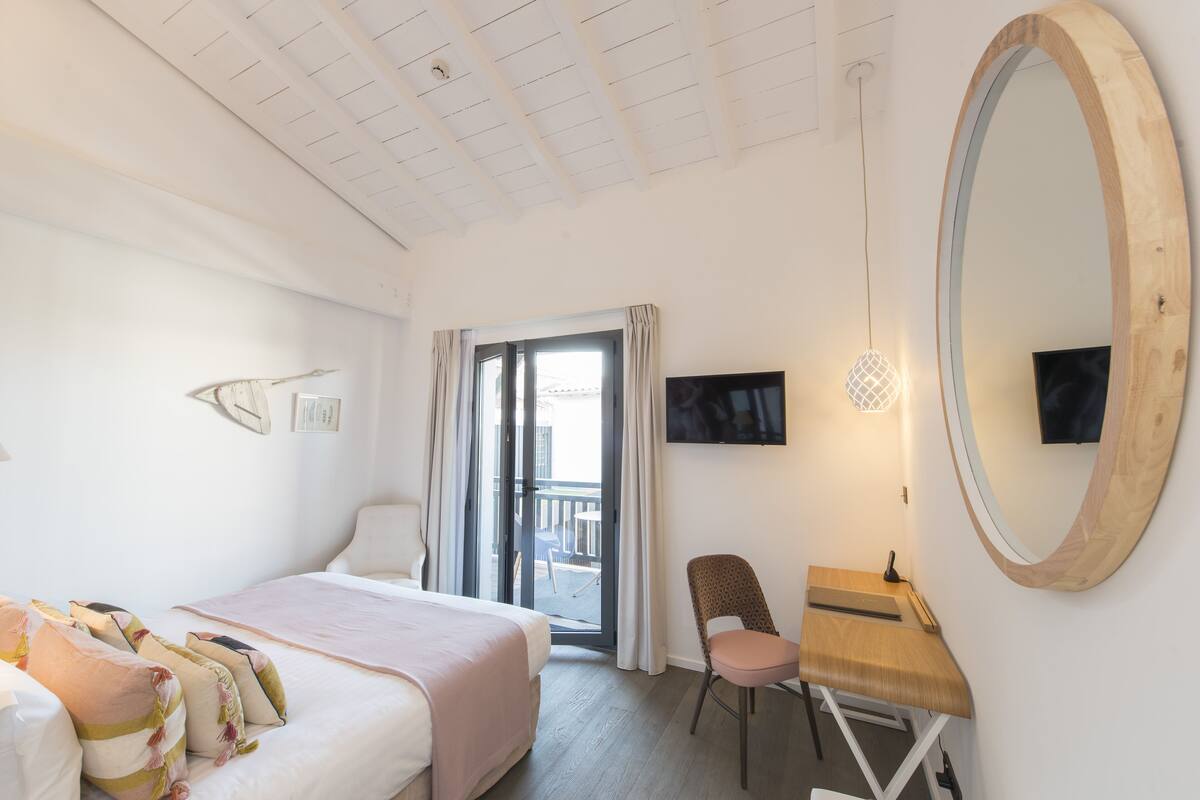 Quarto de hotel com cama, escrivaninha com cadeira, televisão, espelho grande e varanda com cortinas.