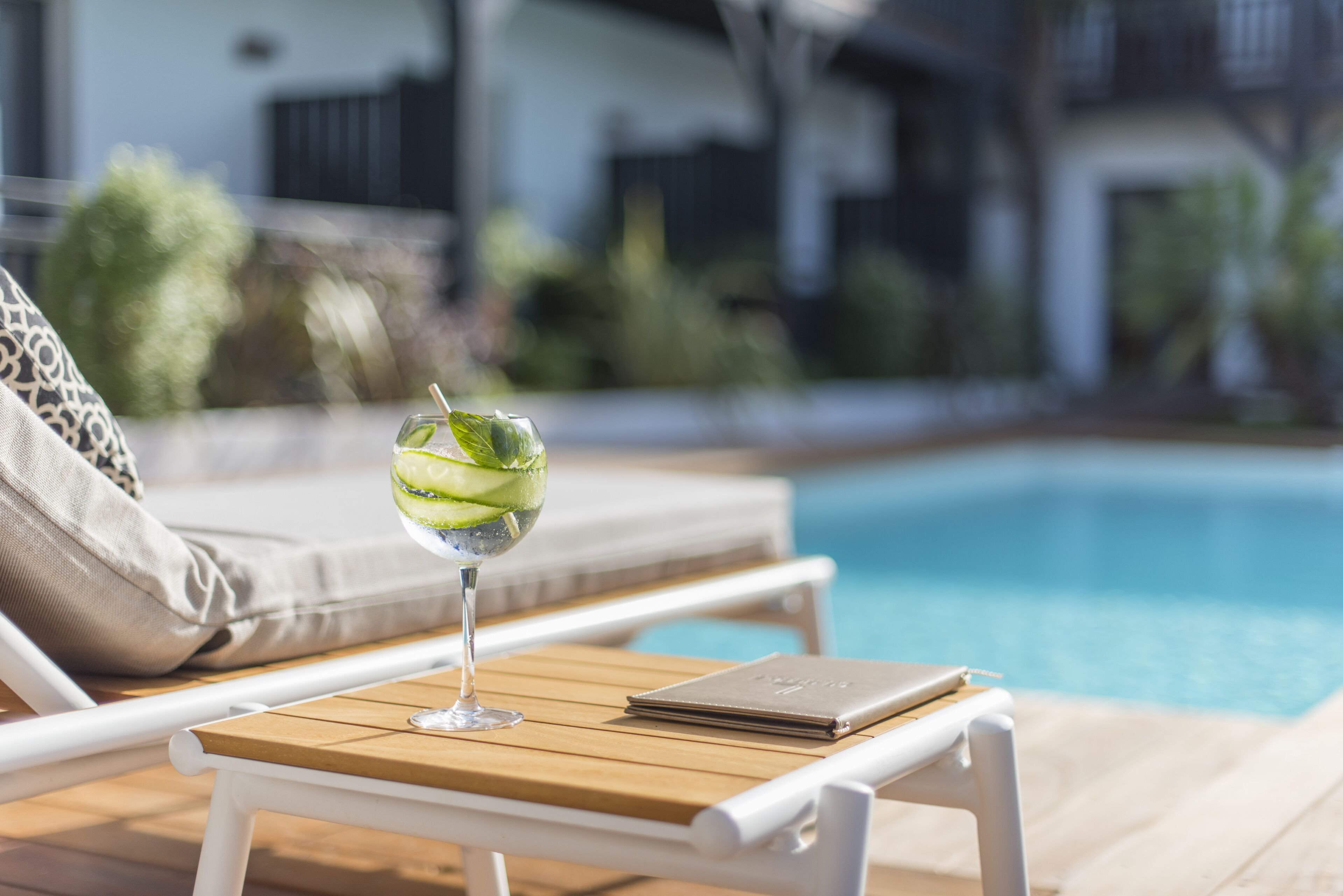 A poolside lounge area with a wooden table, a glass of iced drink, and a notebook.