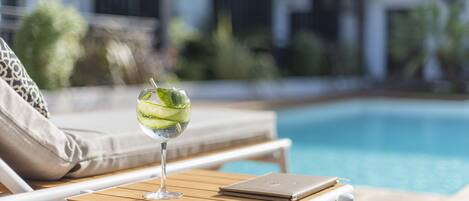 A poolside lounge area with a wooden table, a glass of iced drink, and a notebook.