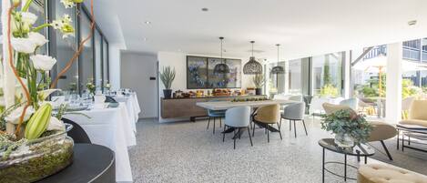 Un comedor moderno con una mesa larga dispuesta para una comida, rodeada de sillas, y una zona de cocina con encimera y plantas.