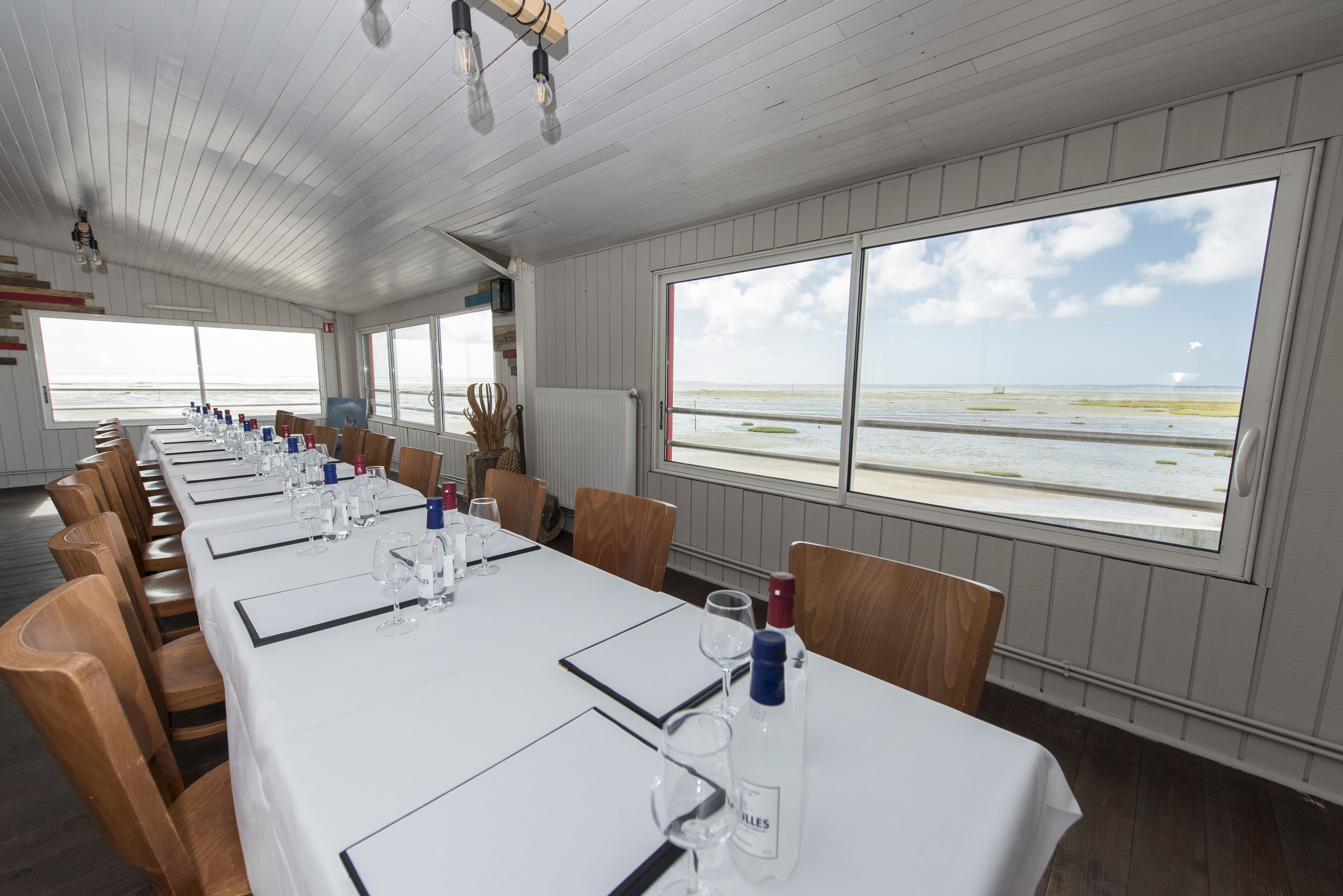 A long dining table set with white tablecloths, wooden chairs, and glassware. Large windows offer a view of the beach.