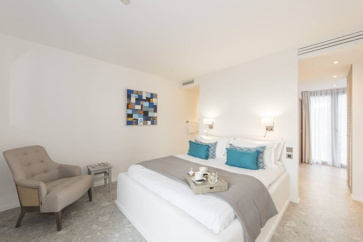 Quarto de hotel moderno com uma cama grande, uma cadeira, uma mesinha e uma obra de arte na parede.
