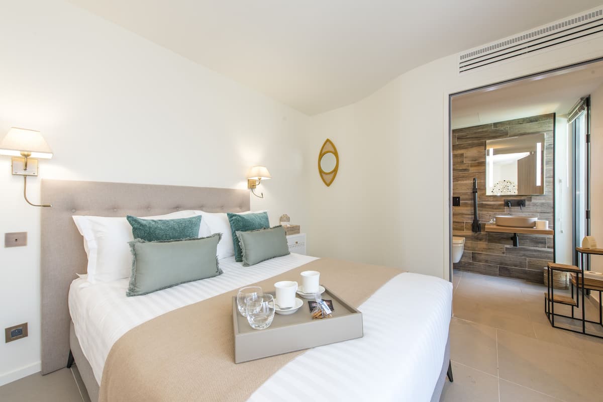 Quarto de hotel moderno com uma cama grande, luminárias laterais, um suporte com copos e lanches, e vista para o banheiro.