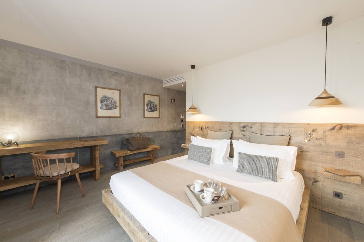 Quarto de hotel moderno com uma cama grande, uma escrivaninha de madeira e uma cadeira. Há duas quadros nas paredes, uma mesinha de cabeceira com uma luminária e um serviço de chá com xícaras e um copo sobre a cama.