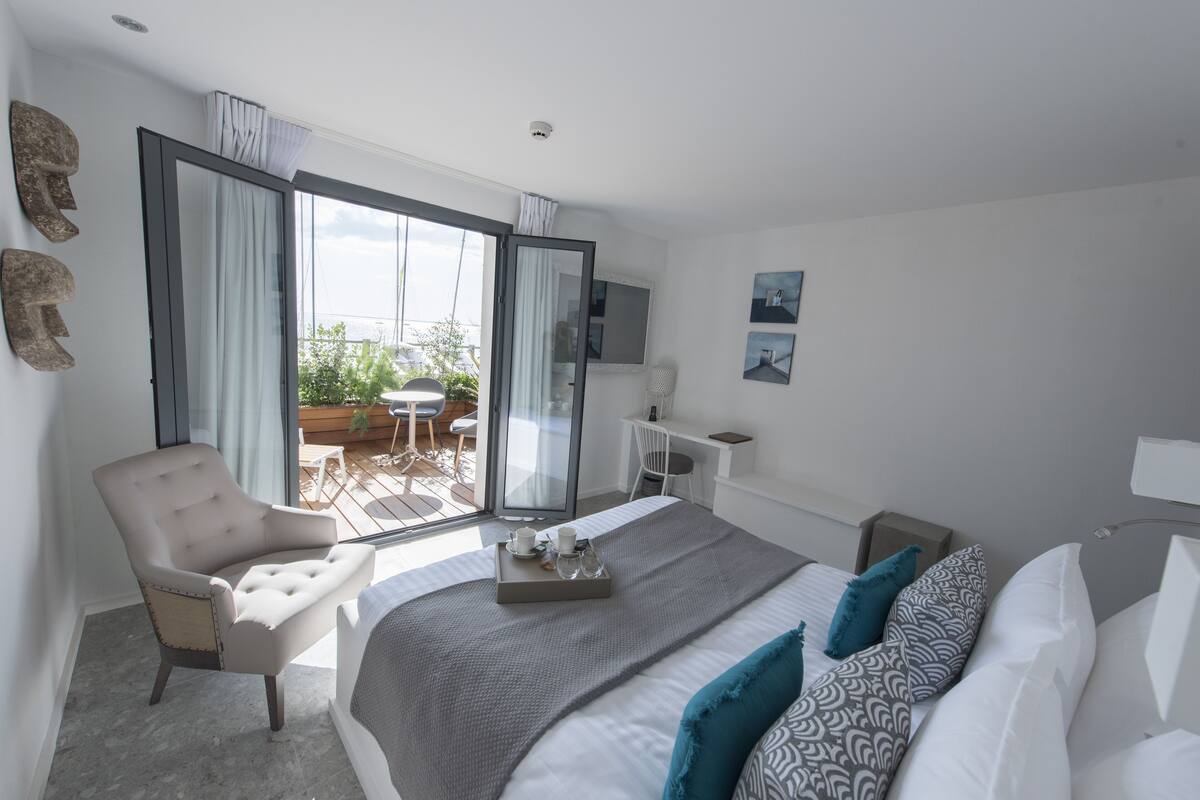 Quarto de hotel moderno com uma cama grande, área de estar com uma poltrona, escrivaninha e varanda com vista para o verde.