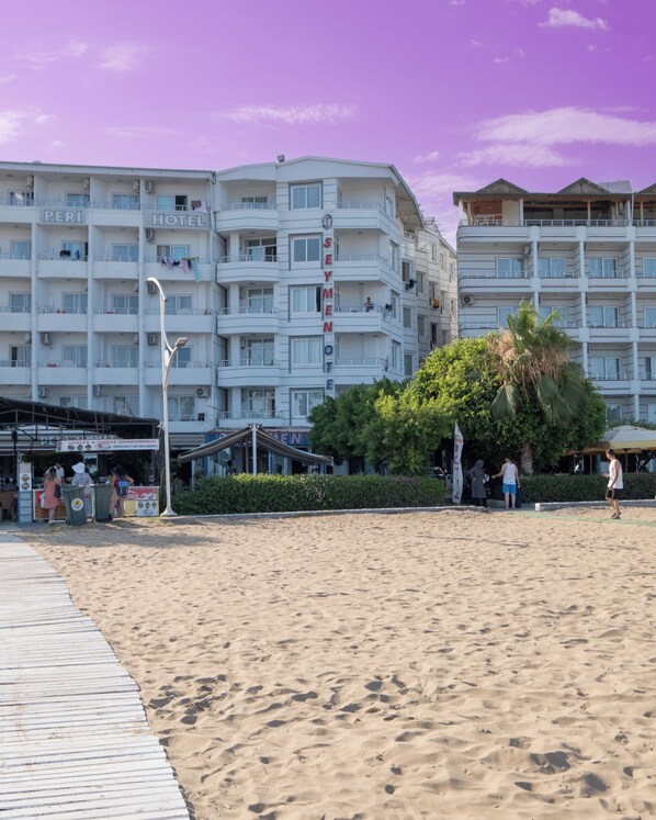 Un hotel frente a la playa con varios balcones, un restaurante a pie de playa y un paseo marítimo arenoso.