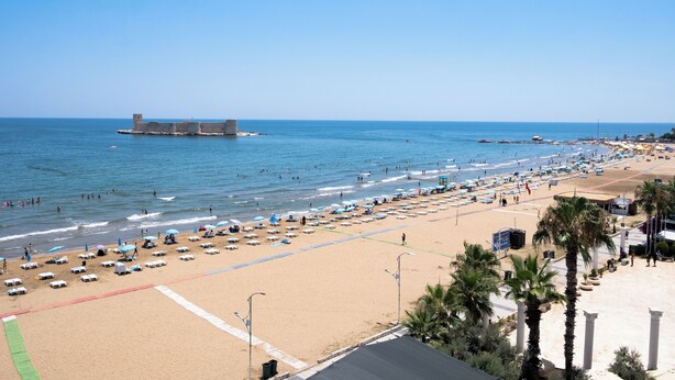 A beach with a large number of umbrellas and sun loungers, a clear sky, and a distant fort-like structure.