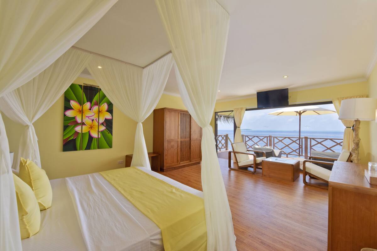 A bedroom with a bed, a painting of flowers, a wooden wardrobe, and a balcony with a view of the sea.