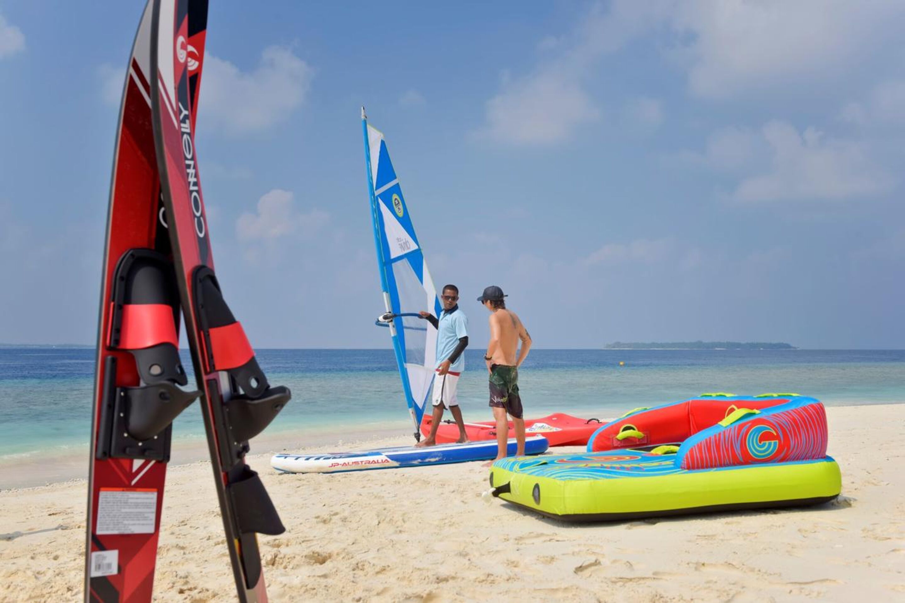 A beach with a windsurfing board, a sail, and inflatable water toys.