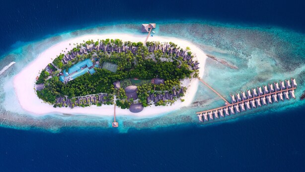 Vista aérea de una isla resort con una piscina central, rodeada de exuberante vegetación y un muelle largo que se extiende hacia el océano.