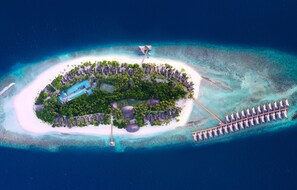 Vue aérienne d’une île-station balnéaire avec une piscine centrale, entourée d’une végétation luxuriante et d’une longue jetée qui s’avance dans l’océan.