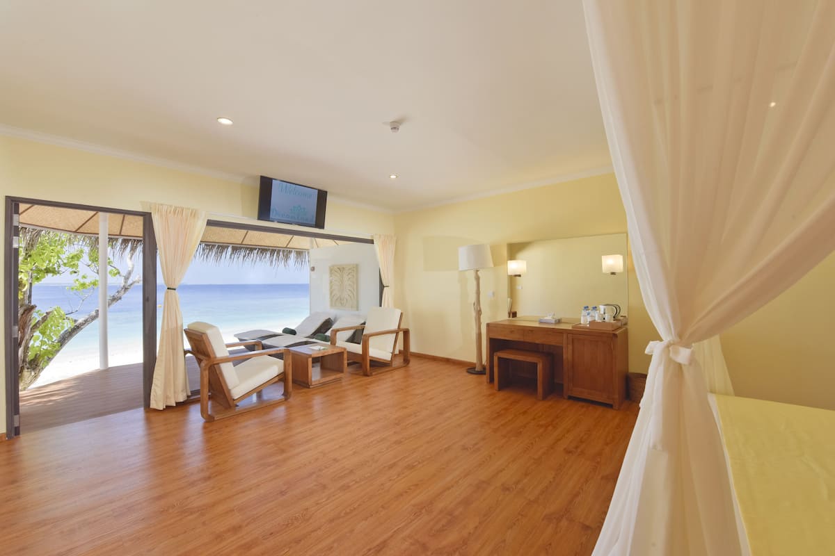 A room with a wooden floor, a desk, two chairs, a bed, a TV, and a view of the sea.