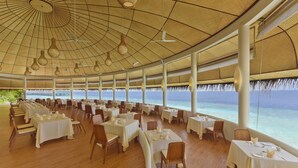 Un restaurante espacioso con mesas dispuestas para comer, ventanas amplias que ofrecen vistas al océano e iluminación colgante en el techo.