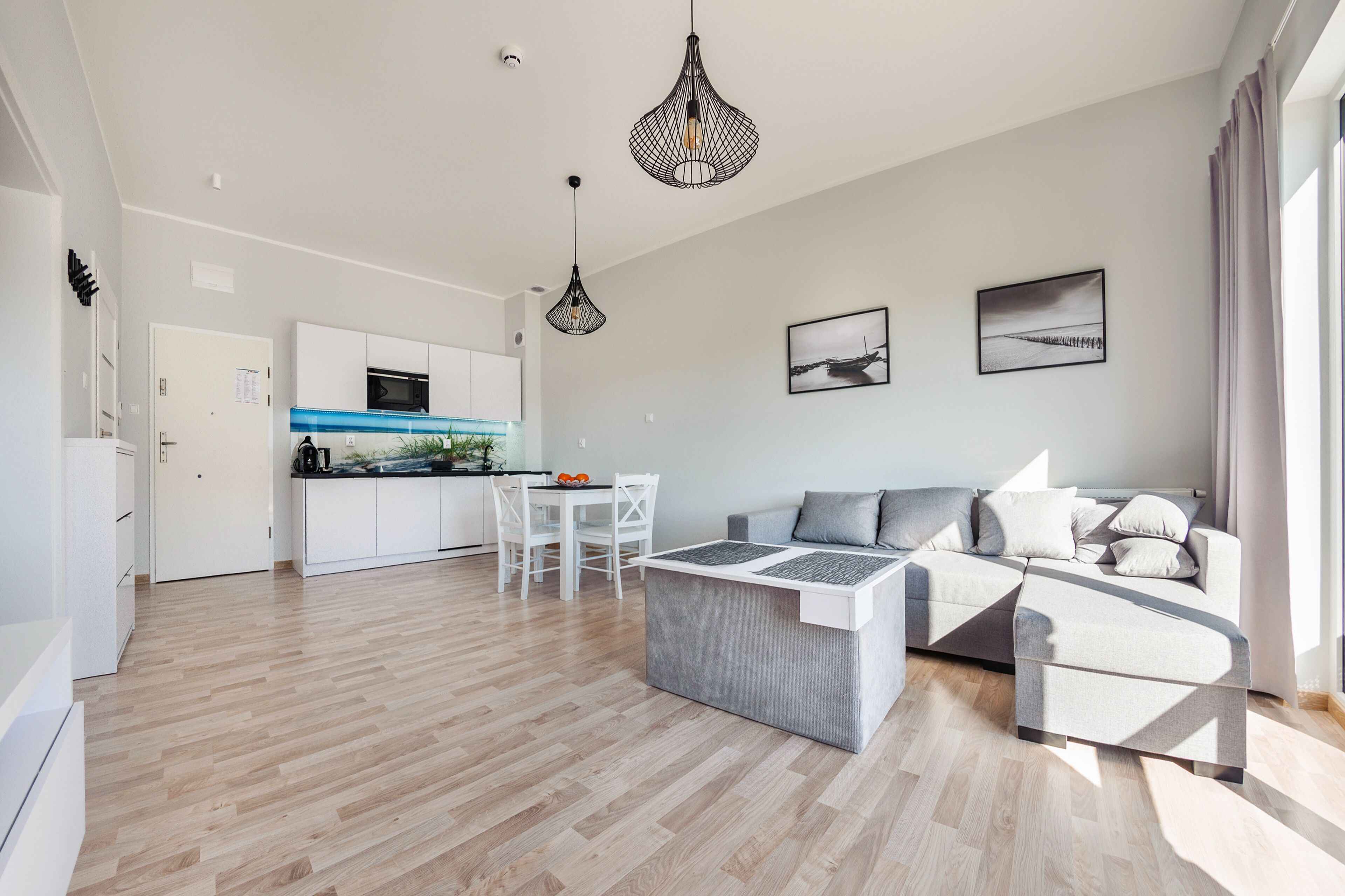 A modern living room with a grey sofa, a coffee table, and a dining area with a white table and chairs. There is a kitchen in the background with white cabinets and a blue backsplash.