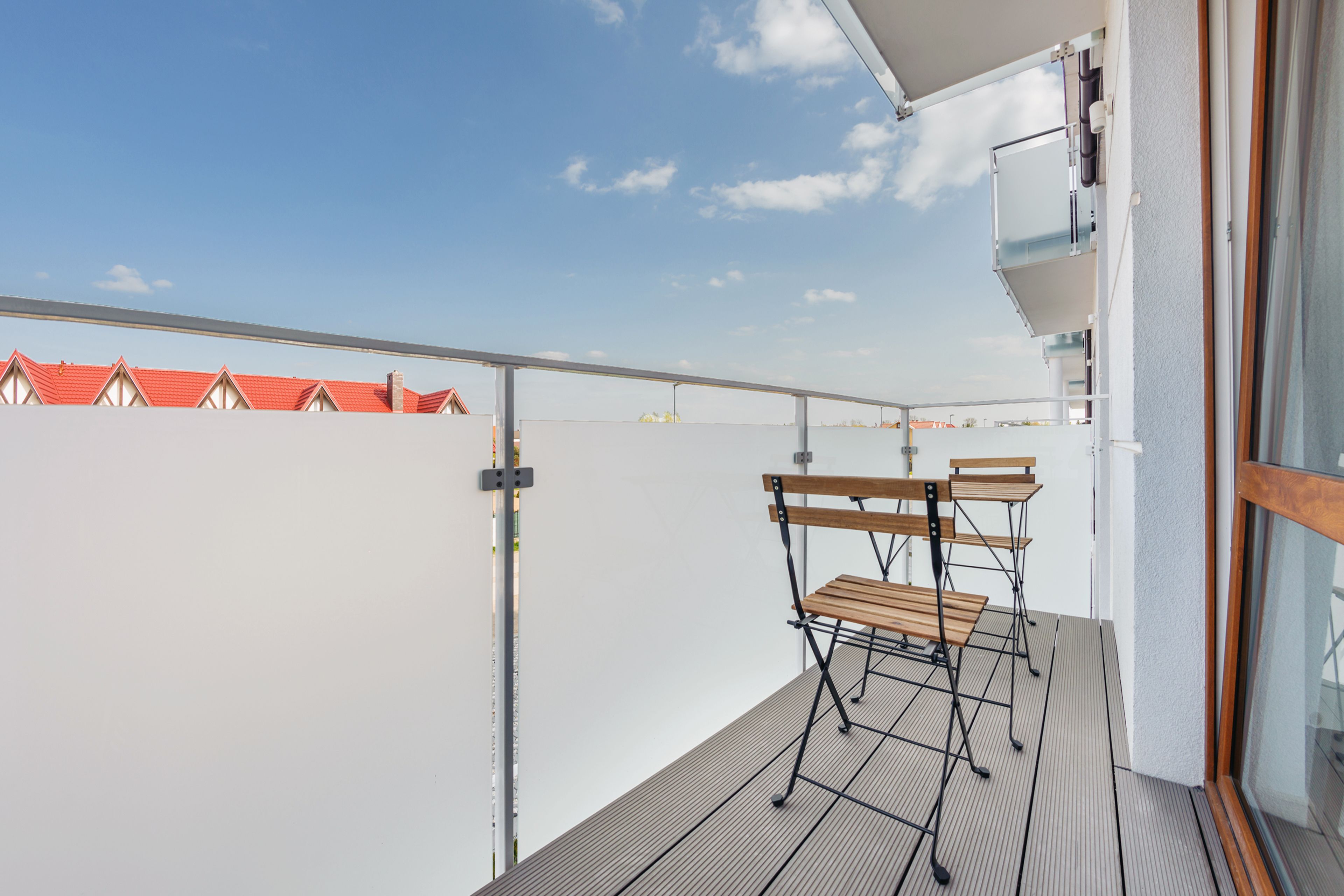 Een balkon met een houten tafel en stoelen, een metalen hekwerk en uitzicht op gebouwen met rode daken.