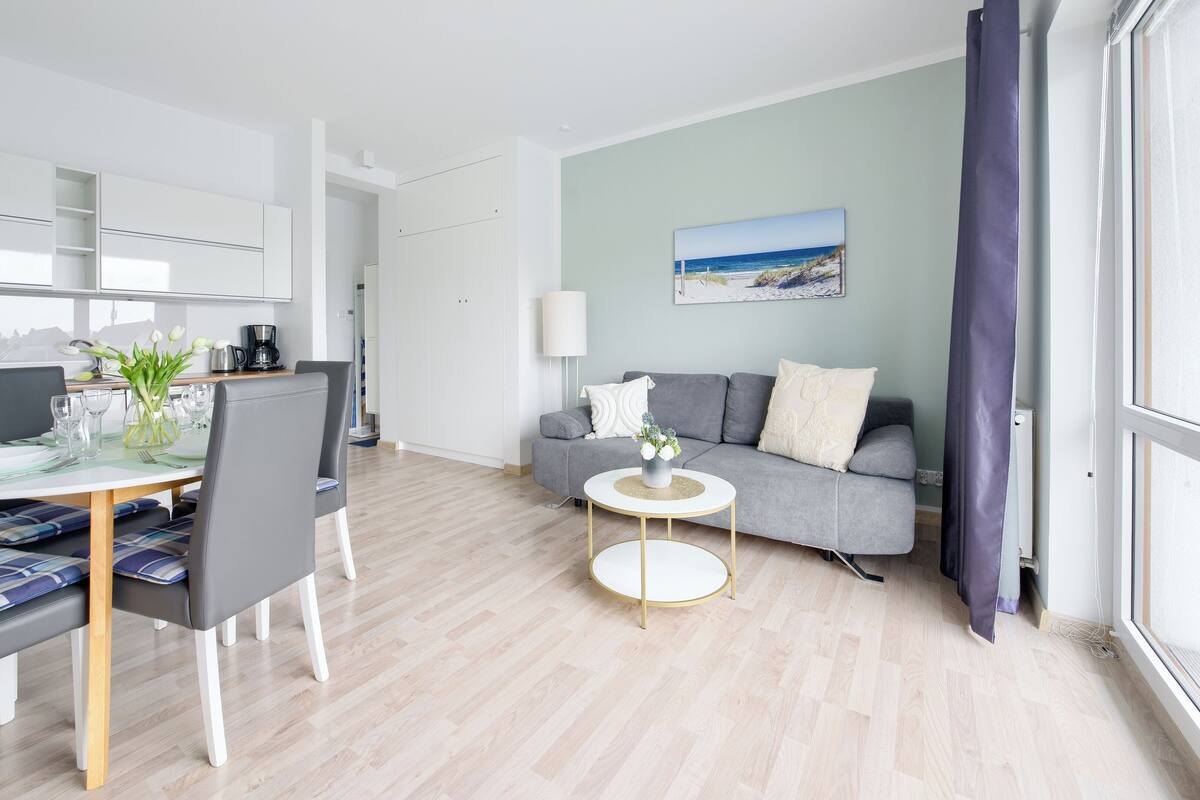 A modern living room with a dining area, a grey sofa, a round coffee table, and a view of the sea through the window.