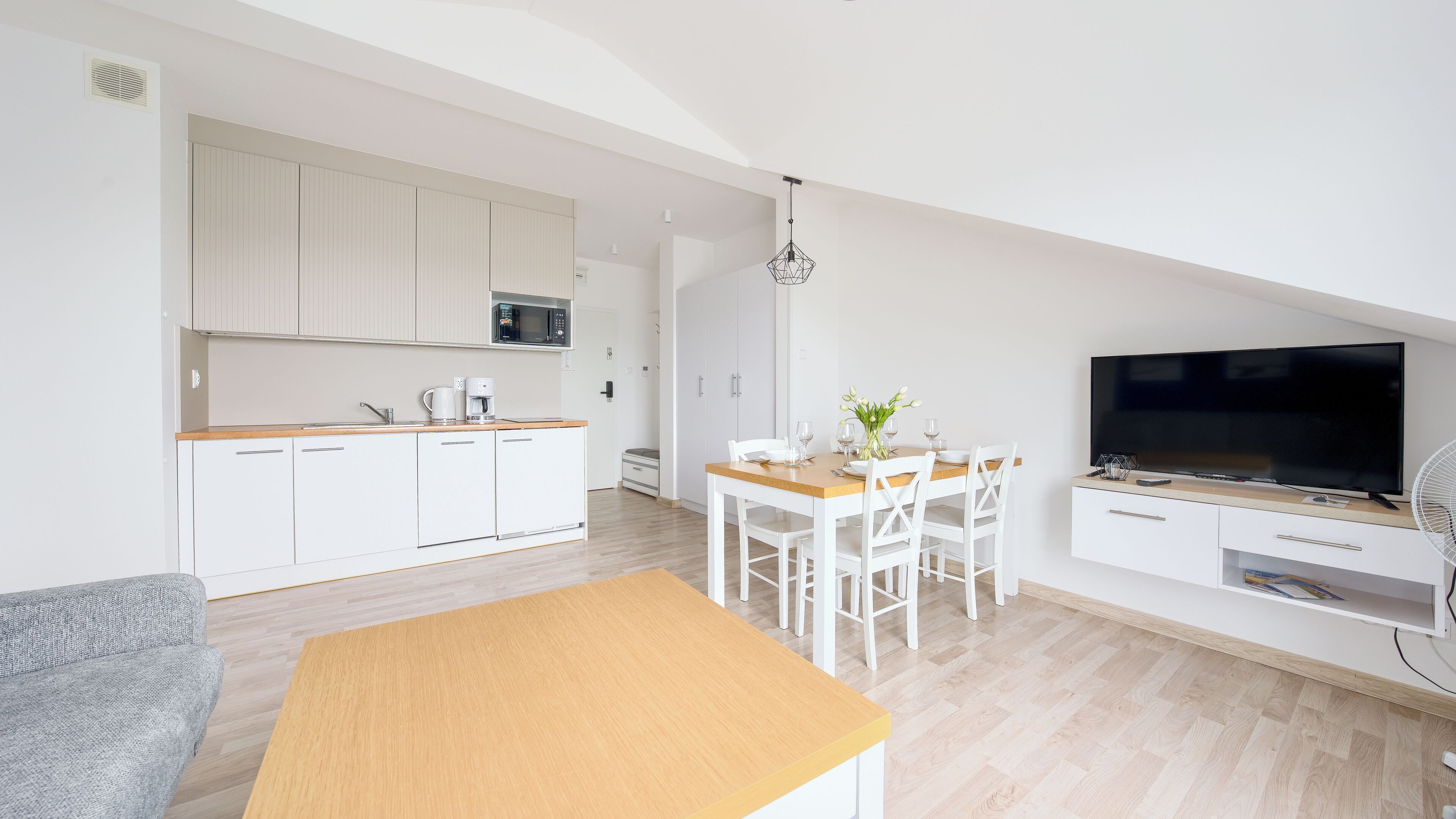 Una cocina moderna con armarios blancos, microondas y cafetera. Hay una zona de comedor con mesa y sillas de madera, y un televisor sobre un mueble.