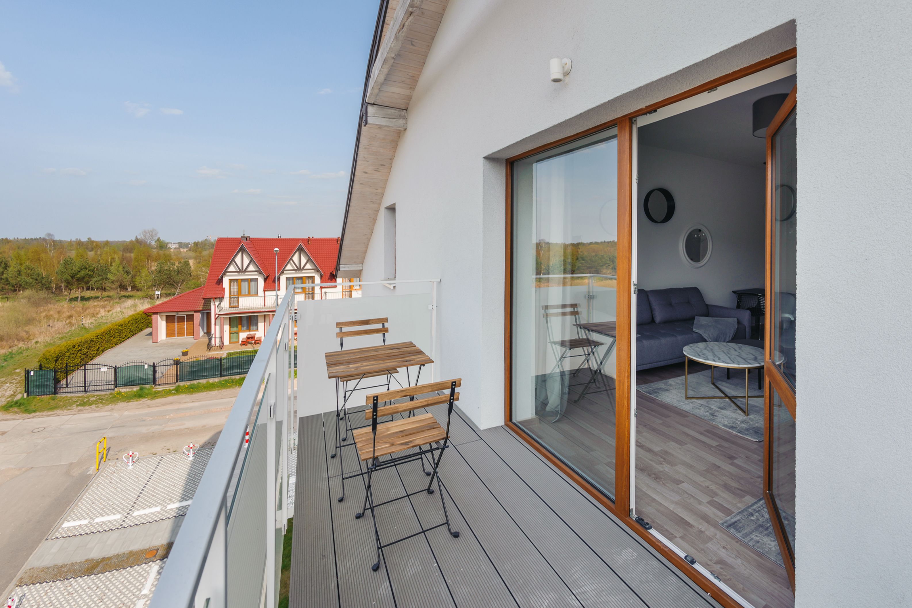 A balcony with wooden furniture, a glass door leading to a room with a sofa and a table, and a view of a red-roofed building in the background.