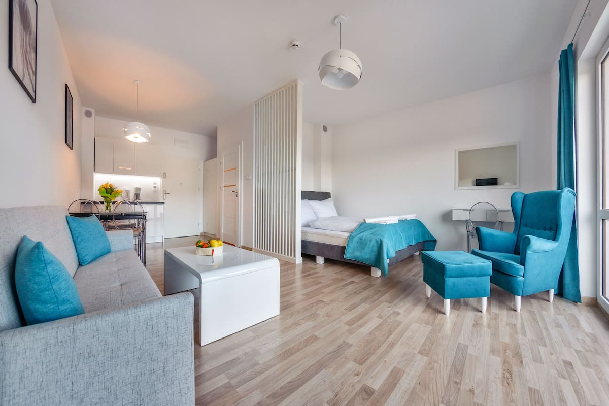 A modern, well-lit room with a grey sofa, a teal armchair, a small white table, and a bed with teal bedding.