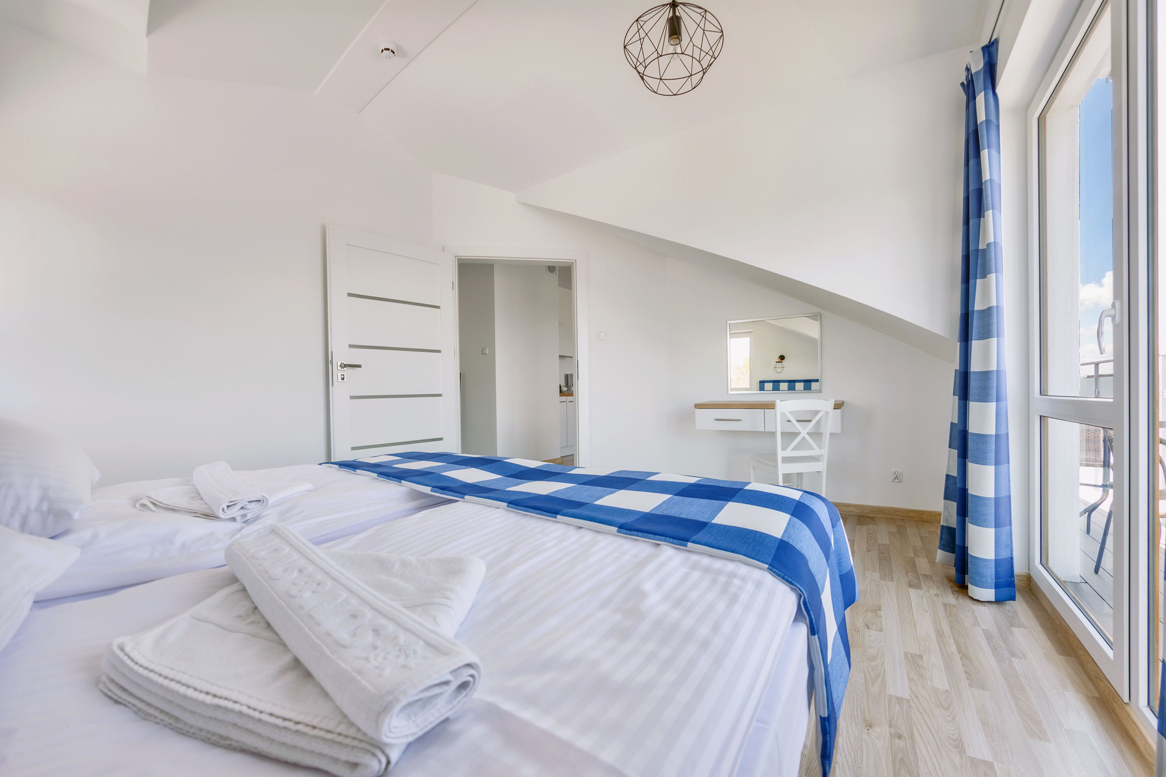Un dormitorio con cama, paredes blancas, una ventana con cortinas azules y un escritorio blanco con espejo.