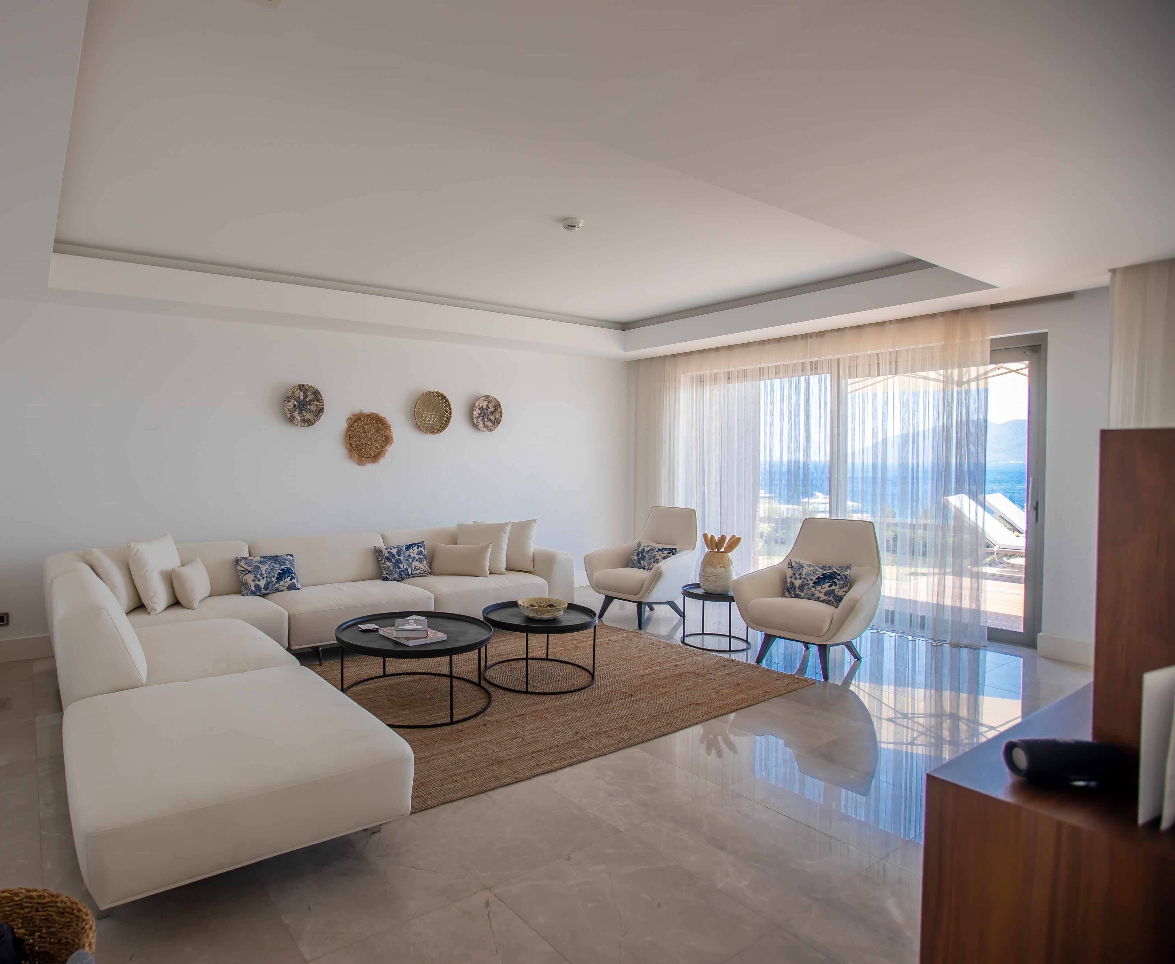 Una sala moderna con un sofá grande, dos sillones, una mesa de centro y vista al mar a través de amplios ventanales.