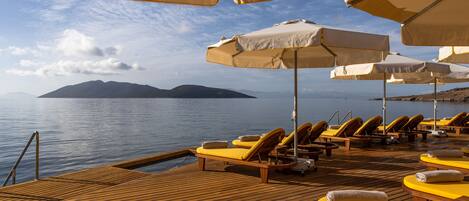 A wooden deck with yellow lounge chairs and white umbrellas overlooking a calm body of water with a distant island.