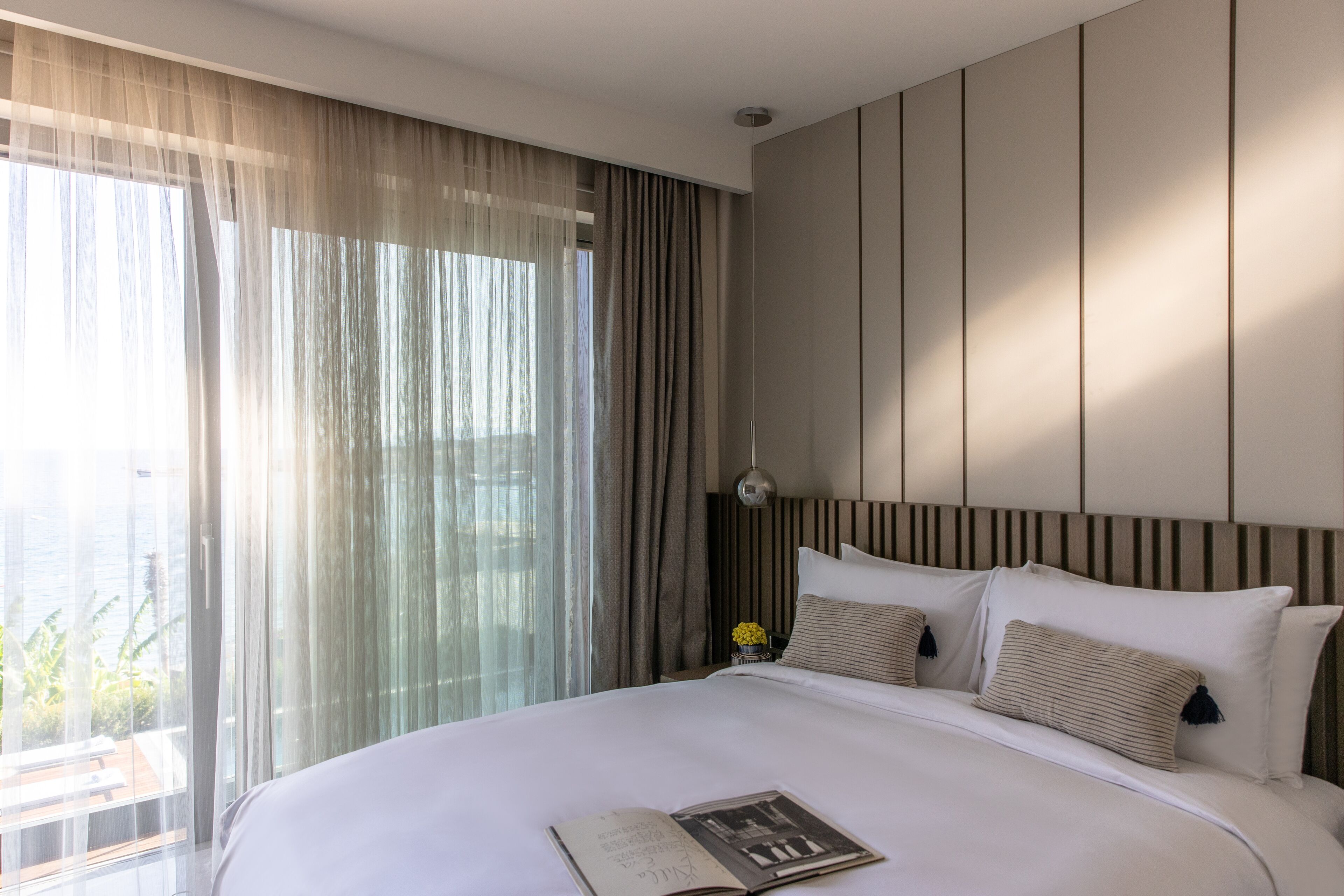 Une chambre d’hôtel moderne avec un grand lit, une table de chevet avec une lampe et des rideaux légers.