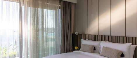 Una habitación de hotel moderna con una cama grande, una mesita de noche con lámpara y cortinas translúcidas.