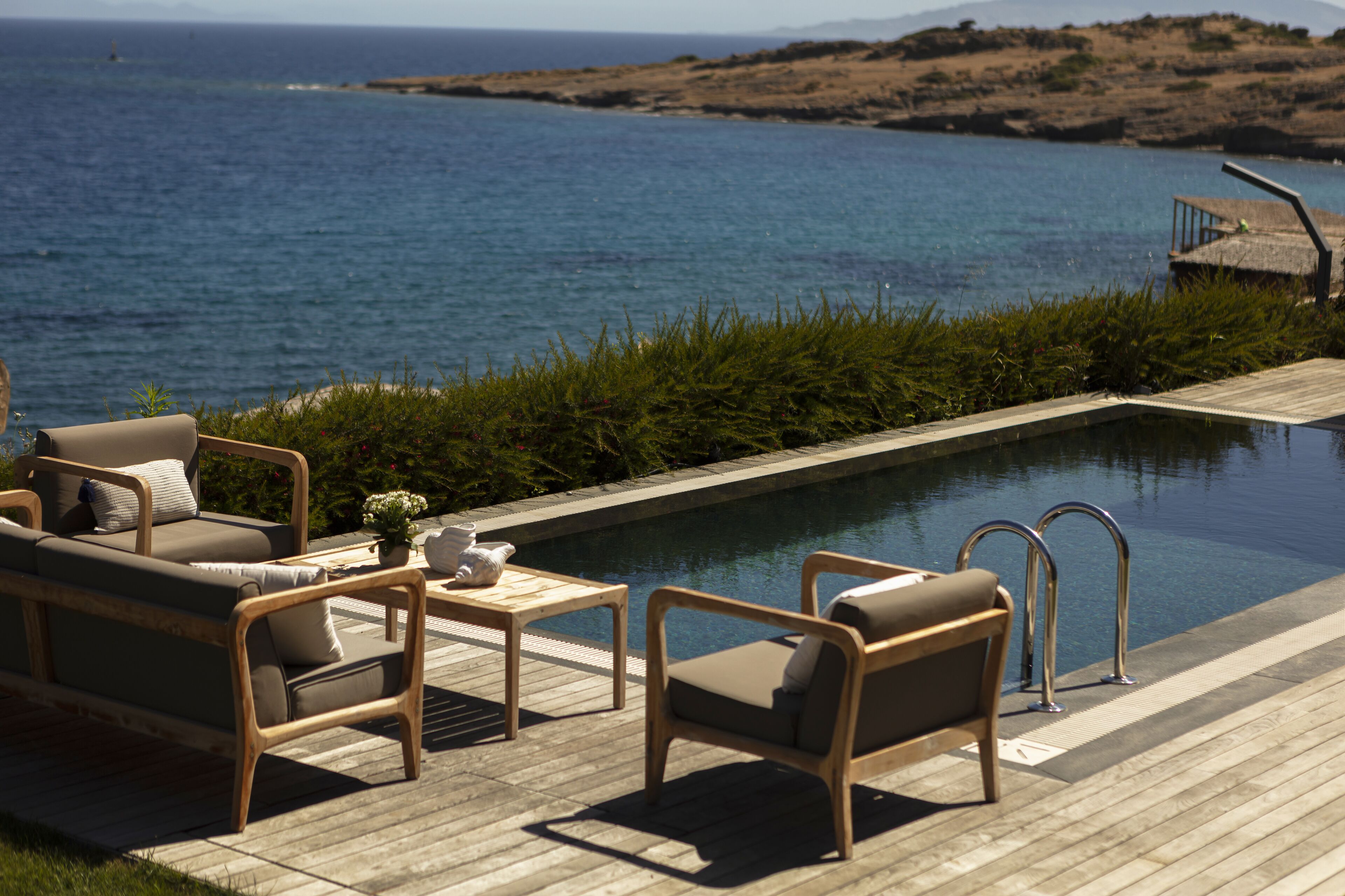 Un área junto a la piscina con cubierta de madera, dos sillones de descanso, una mesita con un florero, y una escalera metálica que lleva a un muelle.