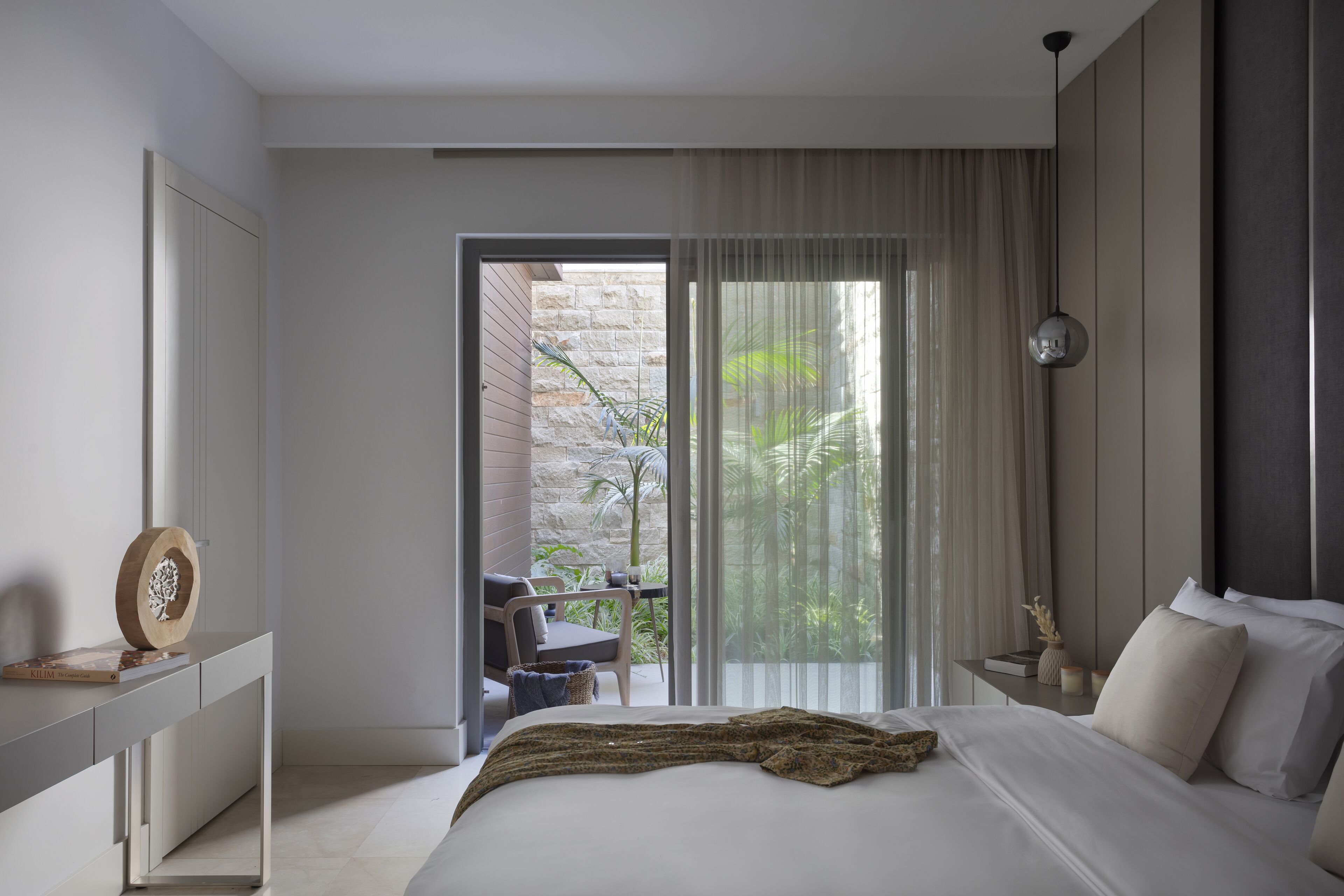A modern bedroom with a large bed, a bedside table with a lamp, a view of a balcony with plants, and a sliding glass door to an outdoor area.