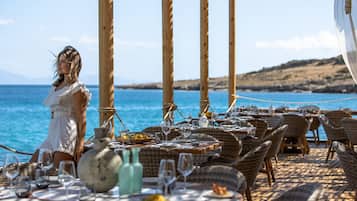 Zona pranzo all'aperto con mobili in vimini e tavoli apparecchiati per un pasto, con vista su uno specchio d'acqua.