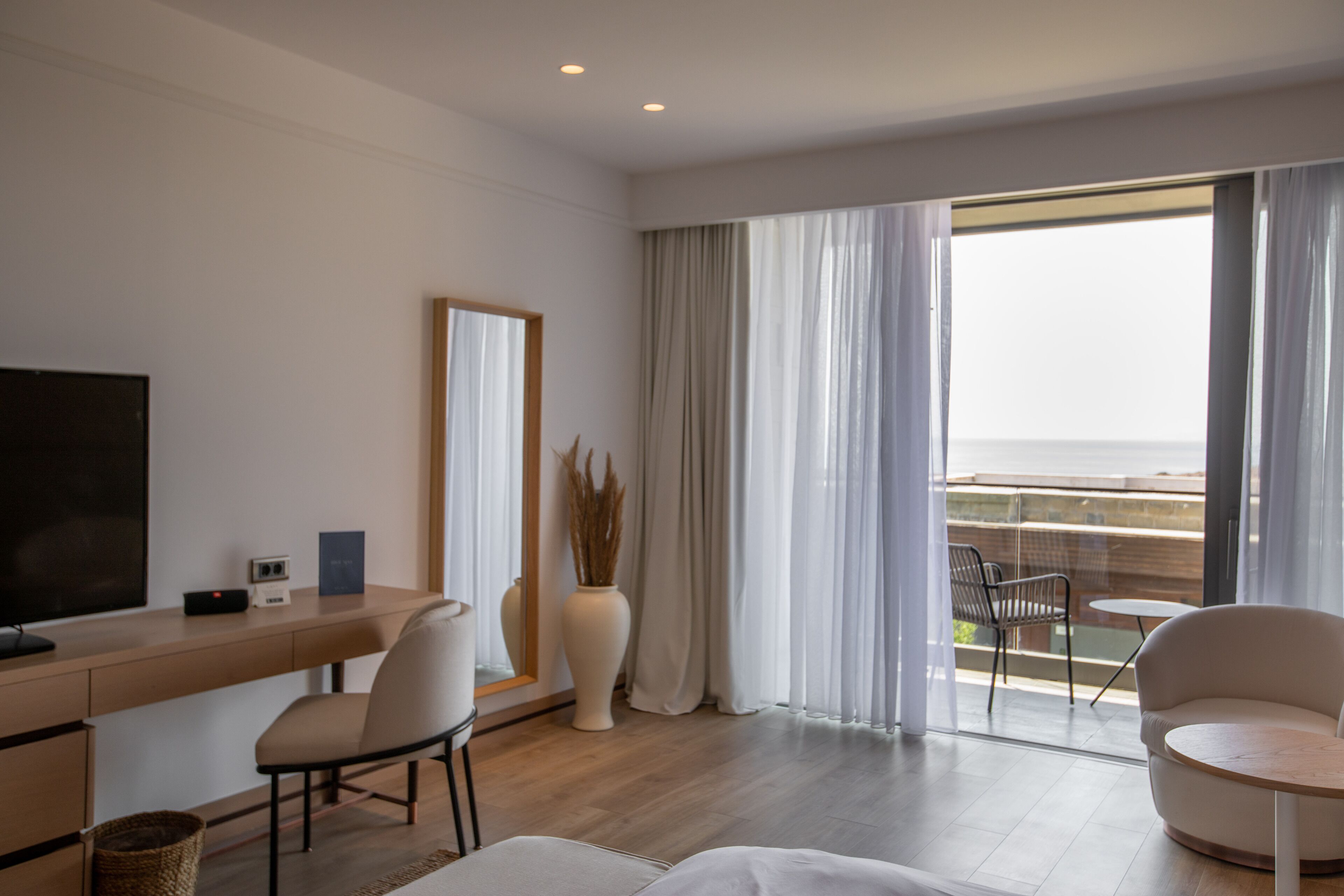 A modern hotel room with a wooden desk, a large mirror, a comfortable chair, a small table, and a view of the sea through large windows.