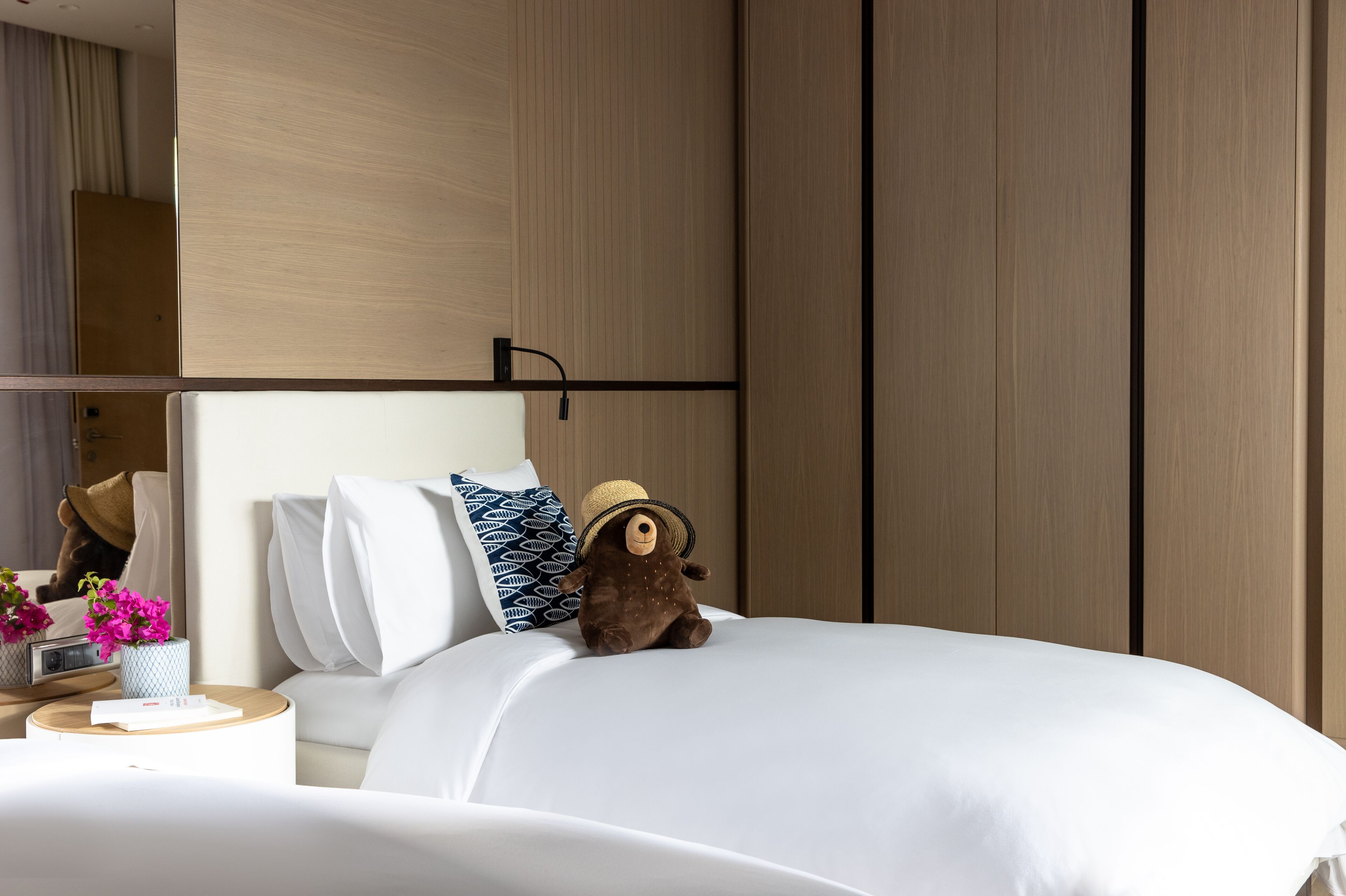 A neatly made bed with a plush toy and a decorative pillow, a bedside table with a vase of flowers, and a wardrobe in the background.