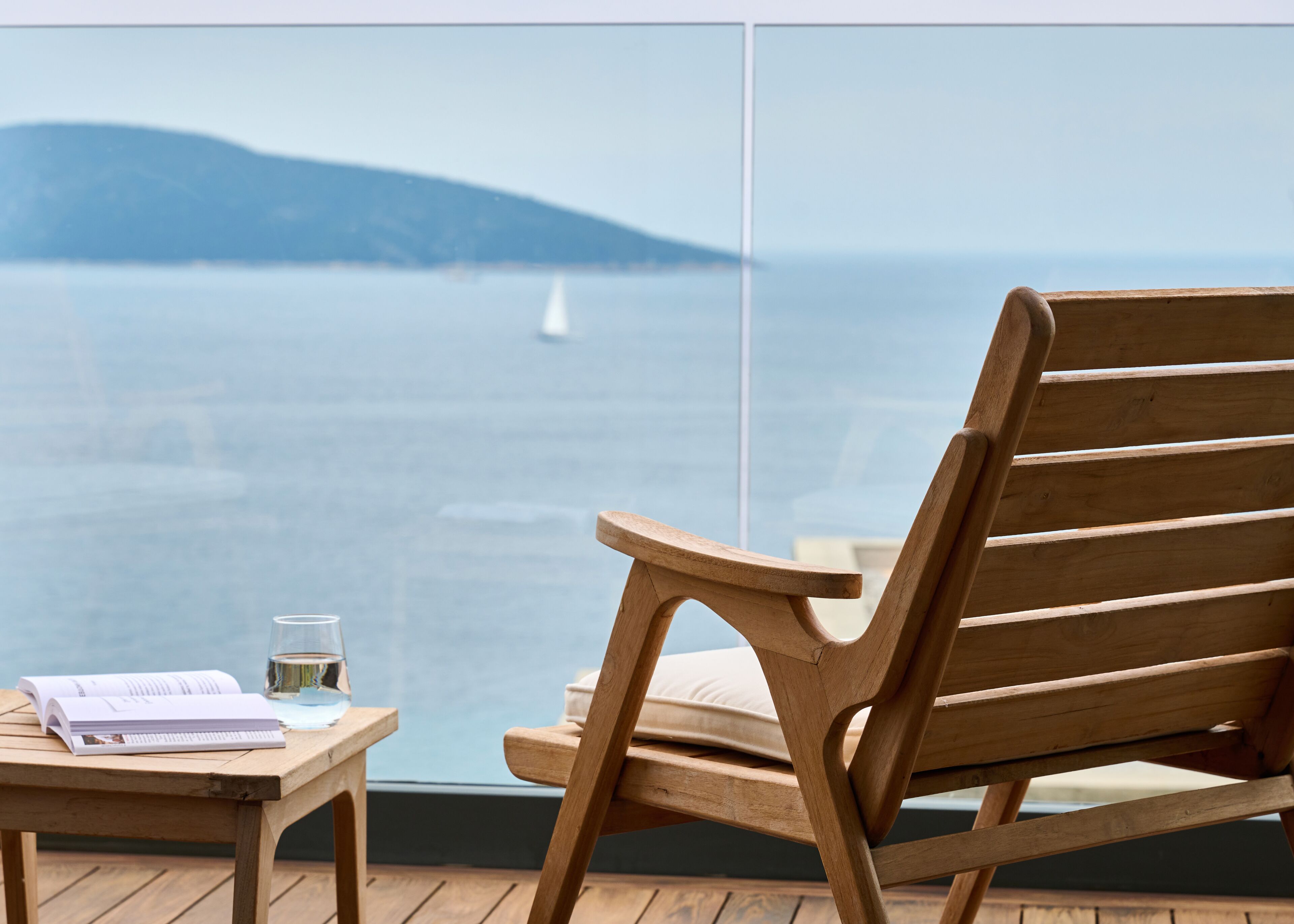 A wooden chair and table on a balcony with a view of the sea and a sailboat.