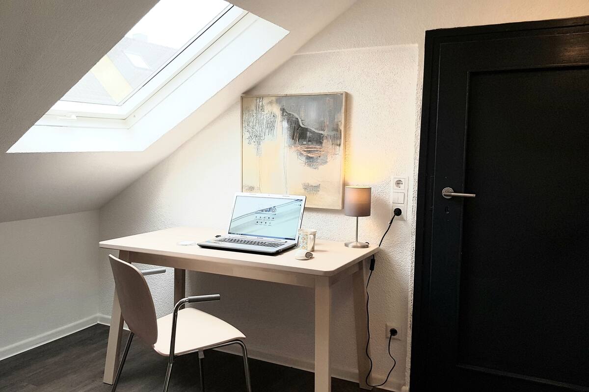 Une petite pièce moderne avec un plafond en pente et un puits de lumière, un bureau avec un ordinateur portable, une chaise et une lampe fixée au mur.
