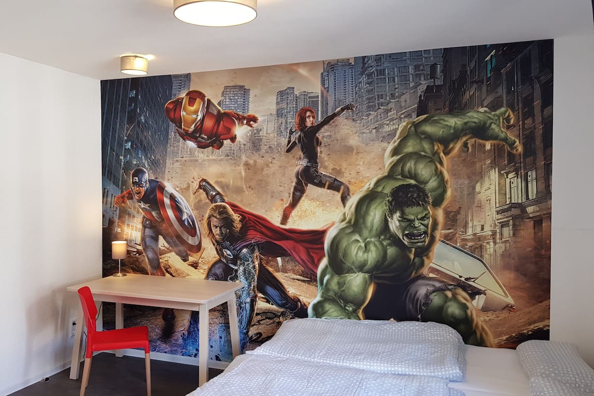 Une chambre d’hôtel avec une grande fresque murale sur le thème des super-héros, un lit, un bureau avec une chaise et une lampe.