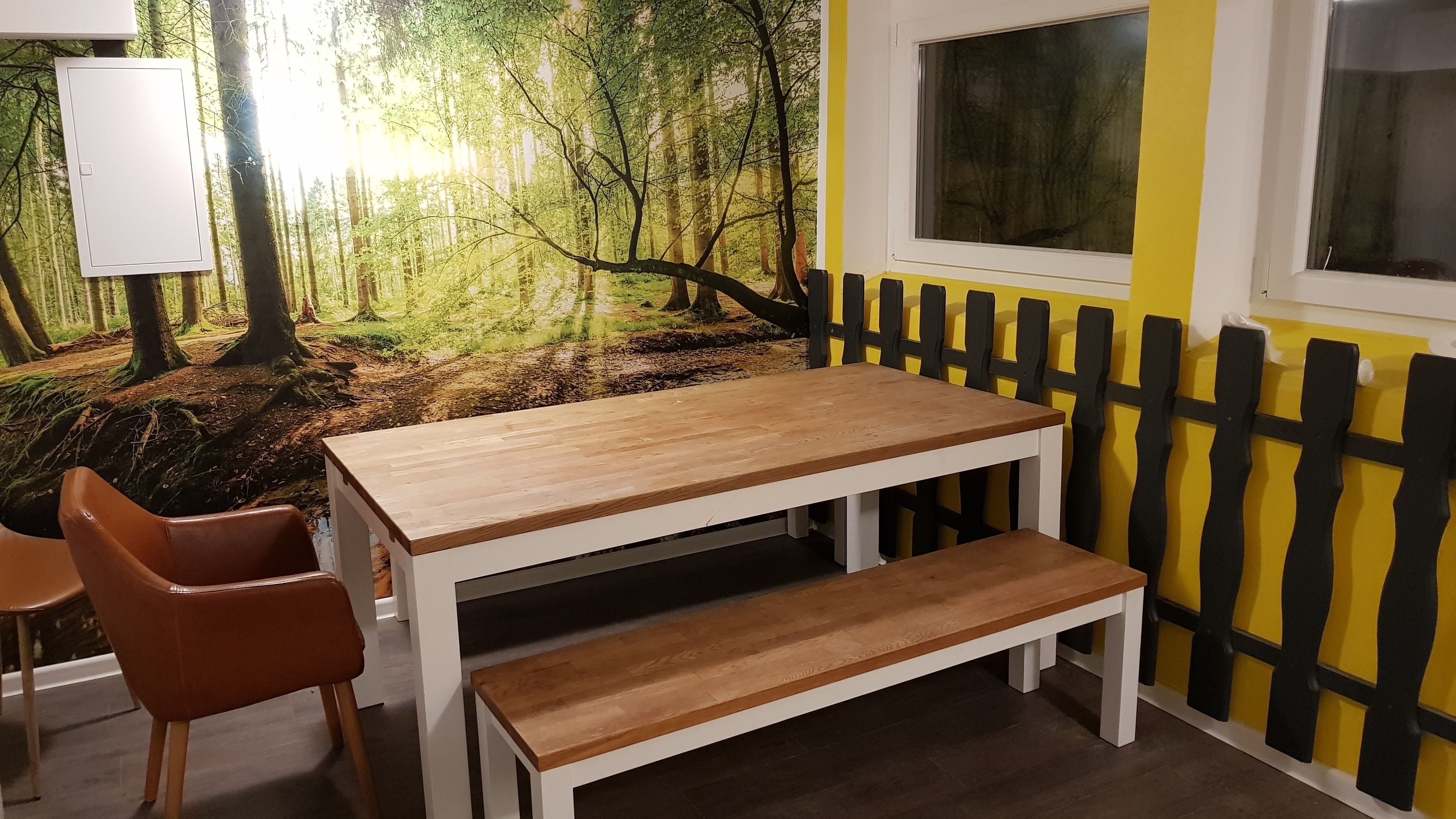 Ein Zimmer mit Holztisch und Bank, einer Tafel und einer gelben Wand mit schwarzem Zaun.