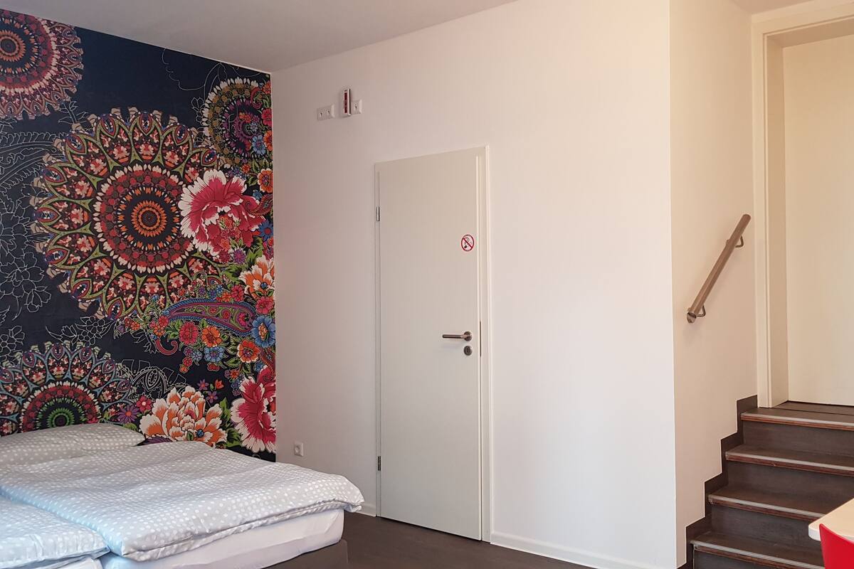 Une chambre à coucher avec un lit, une tapisserie colorée, une porte blanche et un escalier.