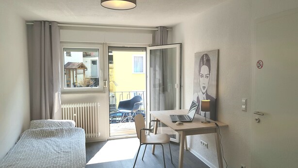 Ein Zimmer mit einem Bett, einem Schreibtisch mit Laptop, einem Sessel und Heizkörper, mit Blick ins Freie.