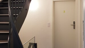 Eine Treppe mit Handlauf, eine Tür mit einer Nummer und ein Rauchmelder an der Decke.