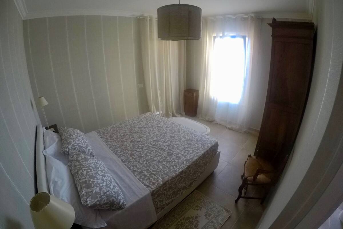 A bedroom with a bed, curtains, a hanging light fixture, and a wooden wardrobe.