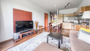 A modern living room with a flat-screen TV, wooden flooring, a dining area with chairs, and a kitchen with wooden cabinets. - Sun & Snow Bałtyk Park (Kolobrzeg)