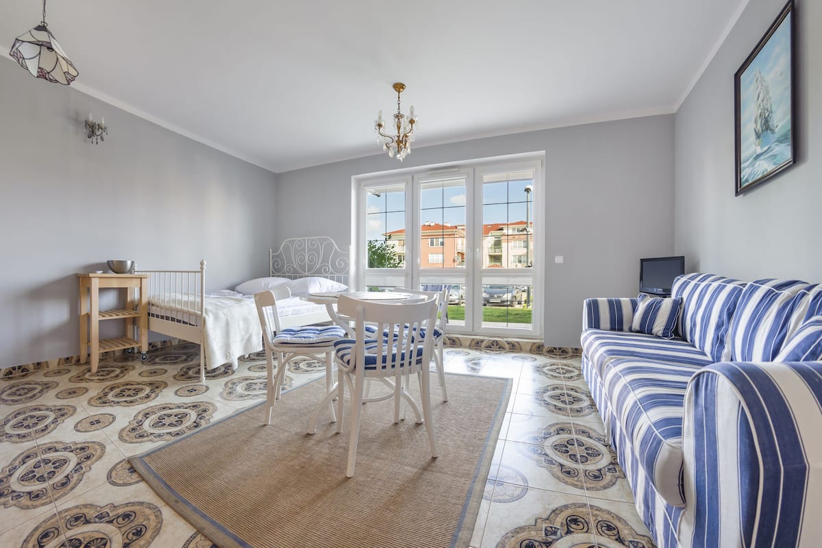 Una habitación con una cama, un sofá a rayas, una mesa de comedor con sillas y un ventanal con vistas a edificios.