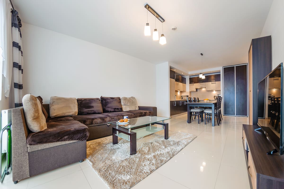 Una sala moderna con una mesa de centro de vidrio, un sofá con cojines, un comedor con mesa y sillas, y una cocina al fondo.