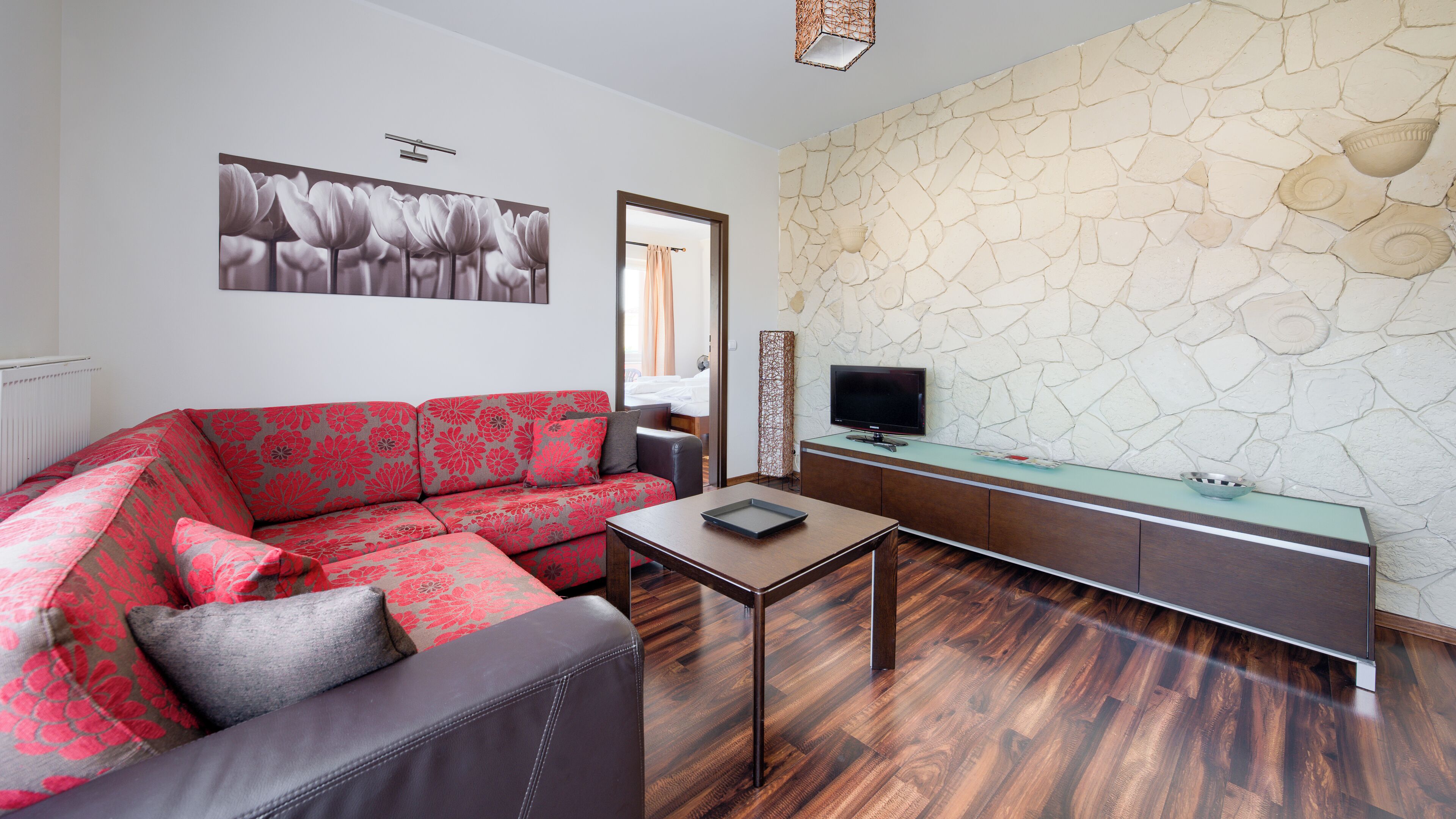 Un salon moderne avec un canapé à motifs floraux rouges, un fauteuil en cuir foncé, une table basse en verre, un téléviseur à écran plat et un mur en pierre.